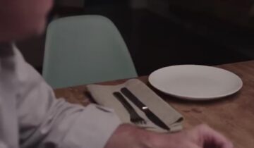 A person sits at a table with an empty plate, napkin, fork, and knife on a wooden surface.
