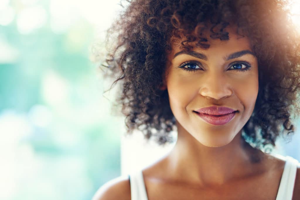 Smiling woman with curly hair in natural light, wearing a white top, with a soft background.